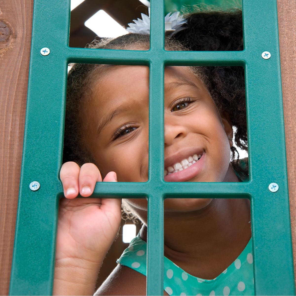 A girl smiling through the green window of a wooden play fort