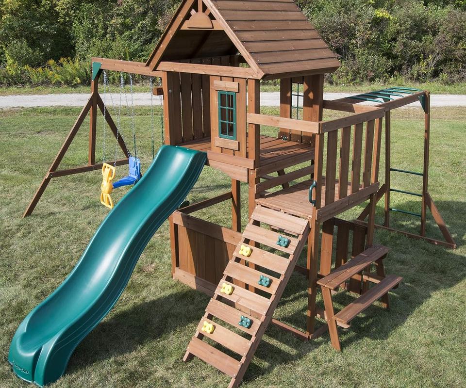 A wooden fort with a shingled roof, two swings and a green wave slide on a lawn